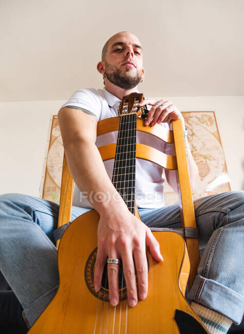 Знизу бородатий концентрований музикант сидить на дерев'яному стільці і тримає гітару вдома вдень — стокове фото