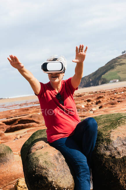 Анонимная пожилая женщина-путешественница, испытывающая виртуальную реальность в очках на берегу моря под облачным небом — стоковое фото