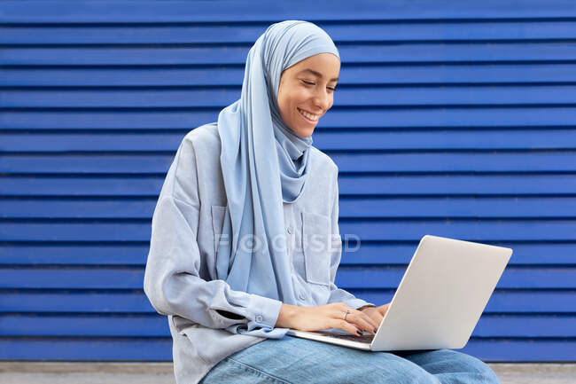 Вміст етнічної жінки в хедкарфі, що друкується на нетбуці, сидячи на синій ребристій стіні на міській вулиці — стокове фото