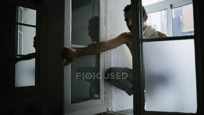 Hombre pensativo sentado de rodillas en el alféizar de la ventana en casa - foto de stock