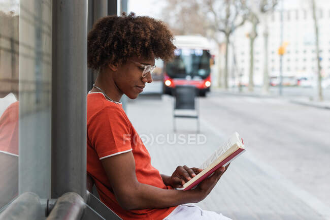 Бічний вид на розслабленого афроамериканця з кучерявим волоссям у повсякденному одязі та окулярах, який читає книжку, очікуючи автобуса на міській вулиці. — стокове фото