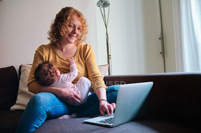 Allegro single madre freelance seduto sul divano e l'allattamento al seno piccolo bambino e la navigazione netbook a casa — Foto stock