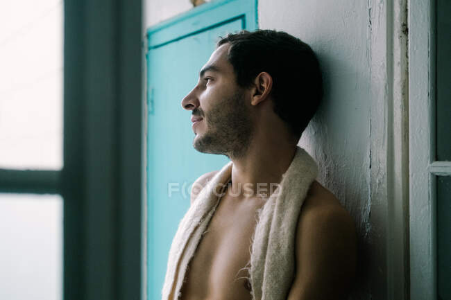 Tranquillo maschio senza camicia con la barba appoggiata sulla parete squallida a casa e guardando altrove — Foto stock