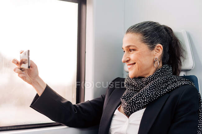 Vista lateral de la hembra de mediana edad positiva con bufanda tomando fotos en el teléfono celular mientras está sentada en el asiento del pasajero cerca de la ventana en el vagón durante el viaje - foto de stock