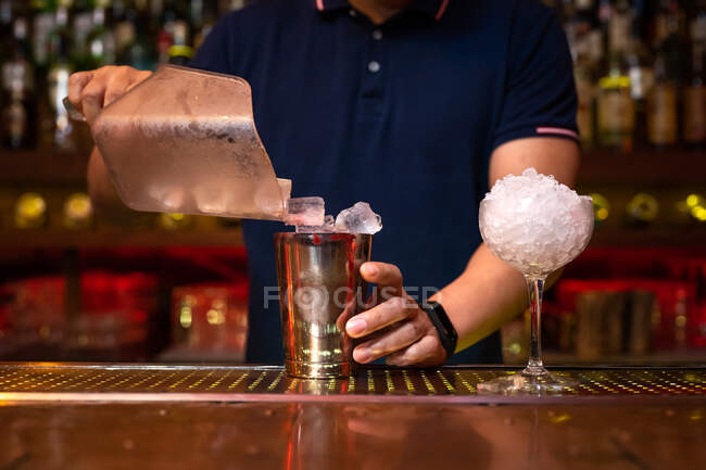 Manos de camarero irreconocible poniendo cubitos de hielo en la coctelera mientras prepara un cóctel en el bar - foto de stock