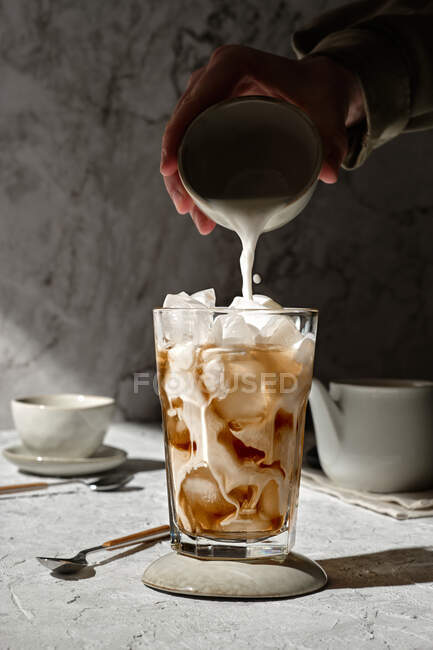 Cultivo persona anónima vertiendo leche de la taza en el vaso con café helado colocado en la mesa a la luz del sol - foto de stock