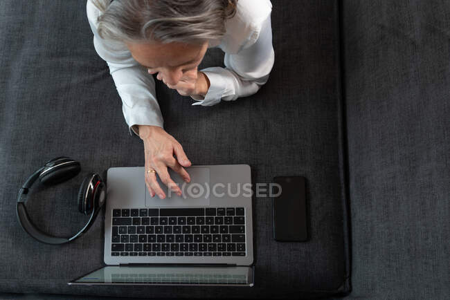Вид сверху анонимной женщины-дистанционного работника, серфингующей по интернету на нетбуке на диване с беспроводной гарнитурой дома — стоковое фото