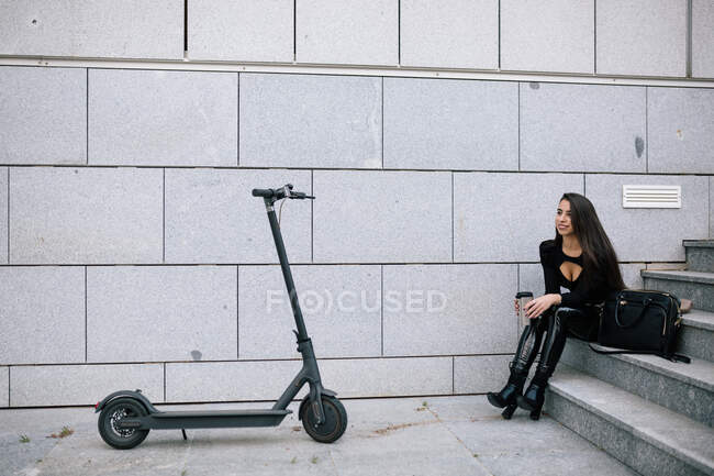 Вид сбоку женщины-предпринимателя, сидящей на лестнице в городе рядом с современным электроскутером — стоковое фото