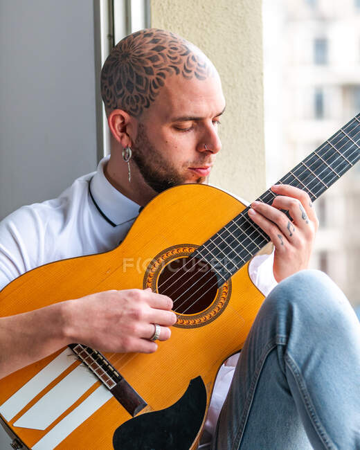 Пенсионер с татуированной лысой головой в повседневной одежде сидит на подоконнике и днем играет на гитаре — стоковое фото