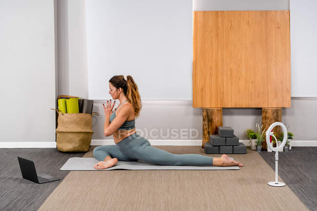 Vista lateral da calma jovem senhora em activewear realizando Half Pigeon ioga pose com mãos de oração durante o treinamento on-line em casa perto de laptop e tripé com smartphone — Fotografia de Stock