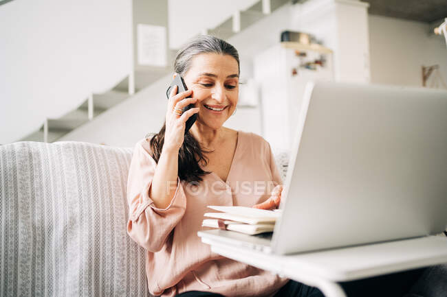Концентрированная женщина средних лет, имеющая телефонный звонок и делающая заметки в блокноте, глядя на экран ноутбука в светлом помещении дома — стоковое фото