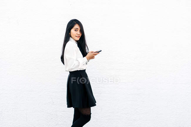 Vista laterale di adolescente etnica femminile in abito formale in piedi vicino al muro bianco e utilizzando il telefono cellulare e guardando la fotocamera — Foto stock