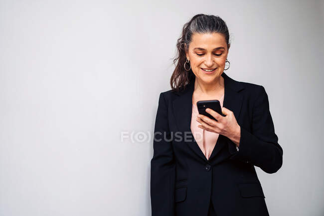 Délice femme entrepreneur d'âge moyen avec queue de cheval portant costume noir de messagerie texte sur téléphone portable tout en se tenant debout sur fond blanc — Photo de stock