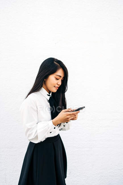 Side view of teen ethnic female in formal outfit standing near white wall and using mobile phone — Stock Photo
