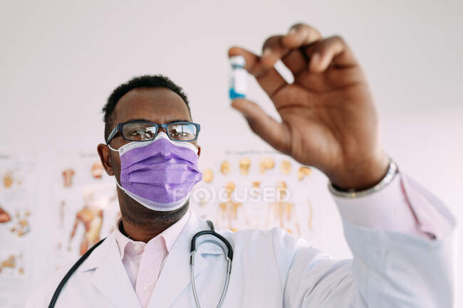 Uomo afroamericano anonimo in uniforme con stetoscopio che mostra vaccino sfocato in mano in ospedale — Foto stock