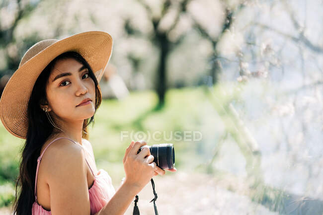 Vue latérale de la femelle ethnique concentrée dans un chapeau de paille prenant des photos à la caméra dans le jardin avec des arbres en fleurs — Photo de stock