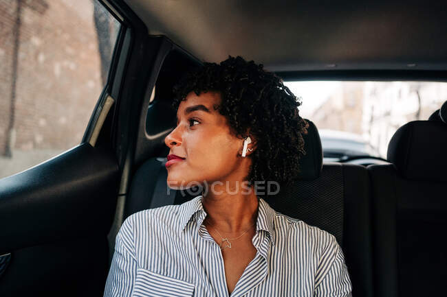 Jeune femme afro-américaine insouciante avec écouteurs TWS écoutant de la musique dans l'automobile moderne regardant par les fenêtres — Photo de stock
