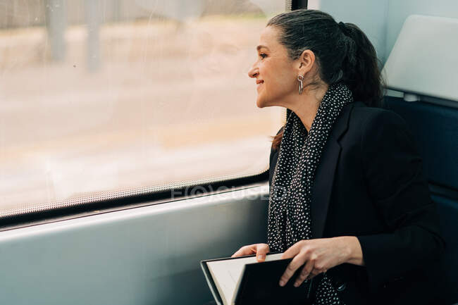 Seitenansicht einer positiven Frau mit Pferdeschwanz, die aus dem Fenster schaut, während sie auf dem Beifahrersitz im Waggon während der Fahrt Buch liest — Stockfoto