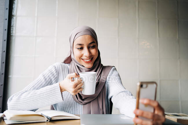 Mulher étnica feliz no hijab e com xícara de bebida tirando selfie no smartphone enquanto desfruta de fim de semana no café — Fotografia de Stock