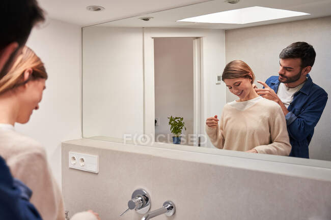 Cultivo enamorado joven hispano macho tocando el pelo de la novia sonriente, mientras que de pie juntos cerca del espejo en el baño en casa - foto de stock