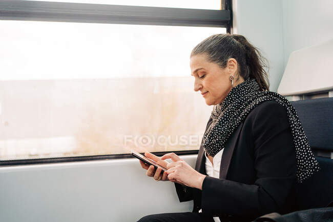 Vista laterale di una femmina positiva di mezza età con sciarpa che prende messaggi sul telefono cellulare mentre è seduta sul sedile del passeggero vicino al finestrino del carro durante il viaggio — Foto stock
