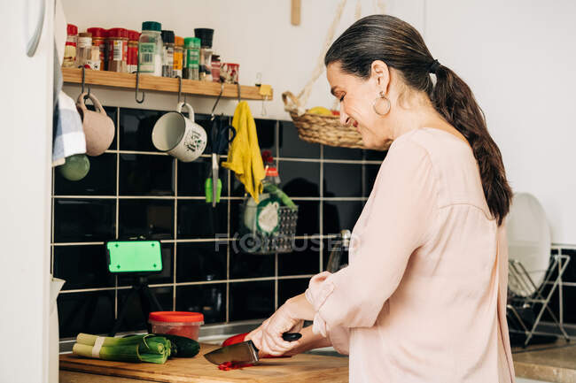 Side view of positive middle aged female with knife cutting red bell pepper on cutting board with leek and cucumbers while cooking in kitchen — Stock Photo