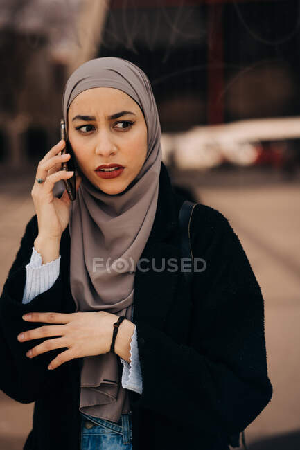 Mujer étnica preocupada en hijab de pie en la calle de la ciudad y tener conversación en el teléfono móvil mientras mira hacia otro lado - foto de stock