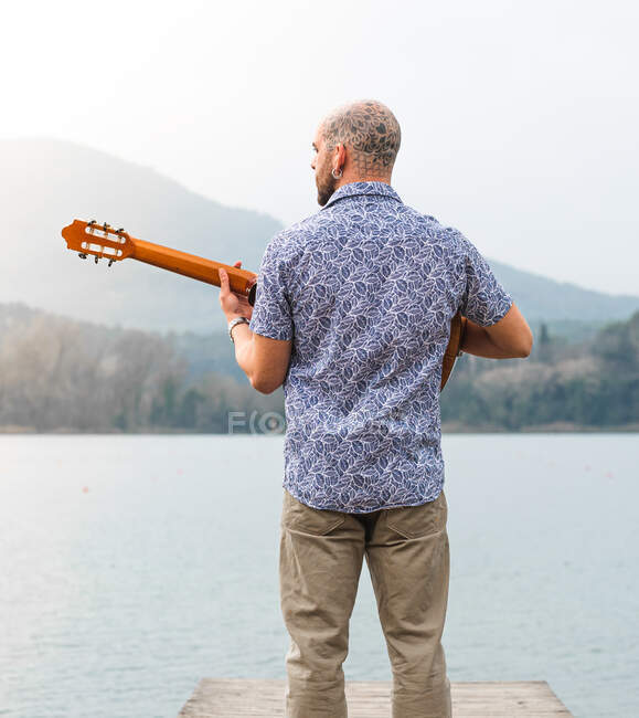 Back view bearded guy in casual clothes standing with guitar on wooden pier near river with mountains on background under cloudy gray sky in daytime — Stock Photo