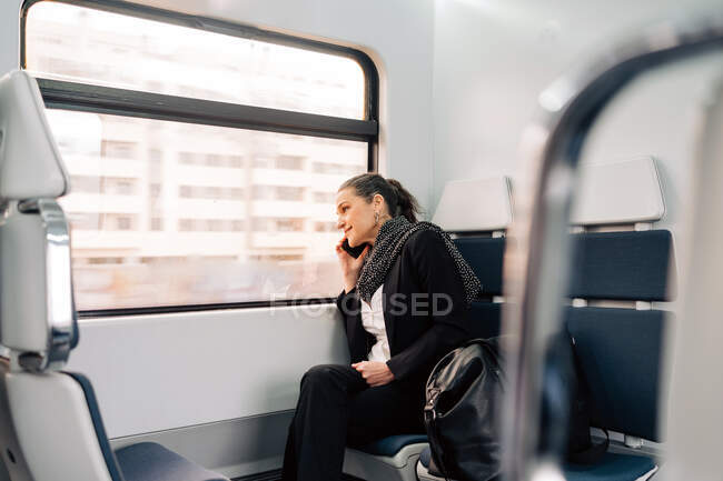 Vista lateral del contenido femenino sentado en el asiento del pasajero y mirando por la ventana mientras tiene llamada telefónica en tren moderno - foto de stock
