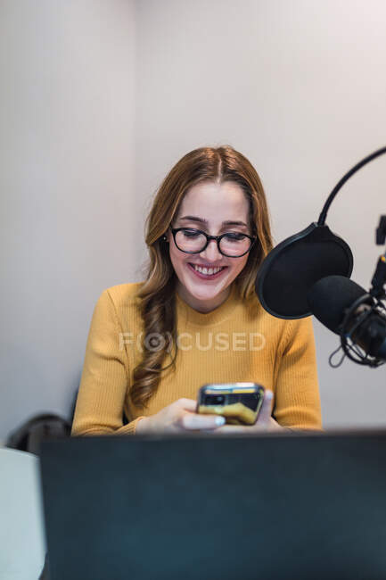 Mujer alegre en anteojos mensajes de texto en el teléfono celular mientras está sentado en la mesa con el ordenador portátil y el micrófono durante el trabajo en el estudio de difusión - foto de stock