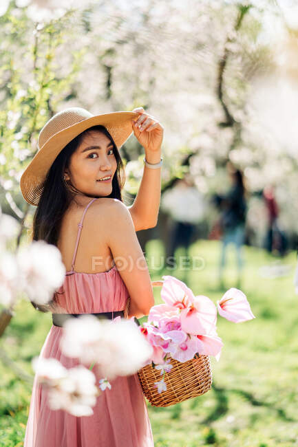 Вид сзади женщины в платье и соломенной шляпе, стоящей с корзиной в цветущем саду, смотрящей через плечо к камере — стоковое фото