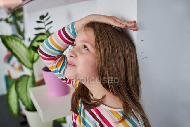 Vista lateral de la alegre linda chica de pie cerca de la pared y medir su altura y mirando hacia arriba - foto de stock