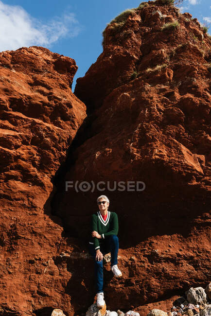 Anziani turista donna in abbigliamento casual seduto su formazione rocciosa con terreno arido sotto cielo nuvoloso blu — Foto stock