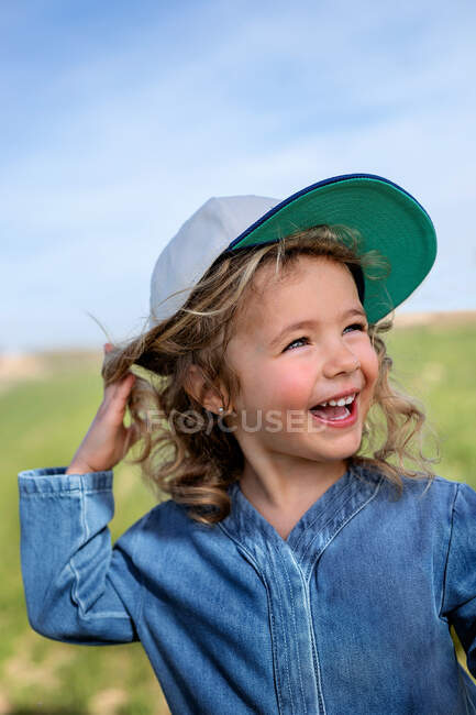 Feliz chica rubia en gorra tocando la cabeza y mirando hacia otro lado contra el cielo azul en verano en el prado - foto de stock