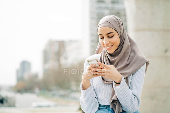 Young ethnic female in hijab standing in city and messaging on mobile phone — Stock Photo