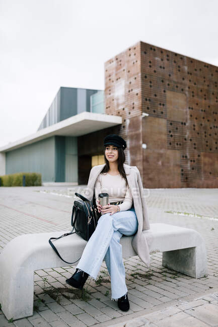 Content female entrepreneur in trendy outfit sitting on bench with cup of beverage to go and looking away — Stock Photo
