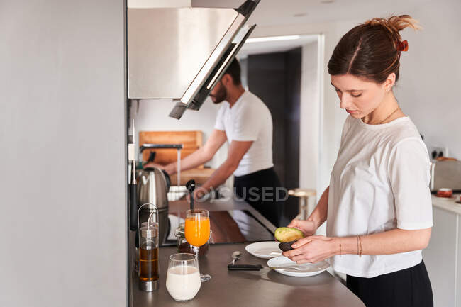 Vista lateral de la tranquila mujer joven en camiseta blanca preparando un desayuno saludable con aguacate mientras está de pie en la mesa en la cocina cerca de novios irreconocibles lavando platos - foto de stock