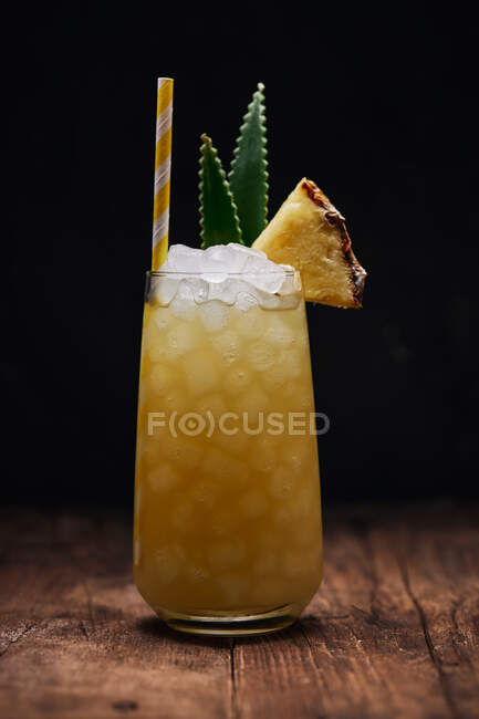 Wooden table with glass of yellow cocktail with ice cubes and refreshing cocktail garnished with spiky leaves and striped straw — Stock Photo