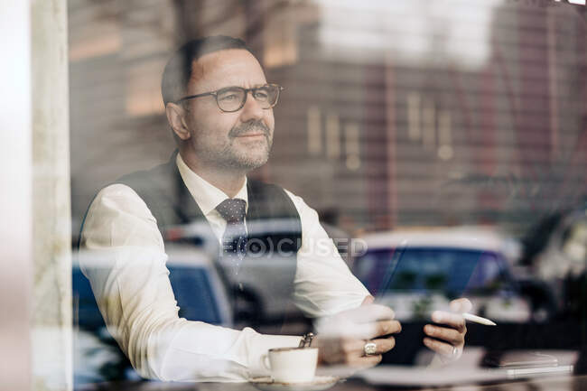 Pensativo empresario masculino étnico de mediana edad utilizando tableta detrás de la pared de vidrio en la cafetería - foto de stock