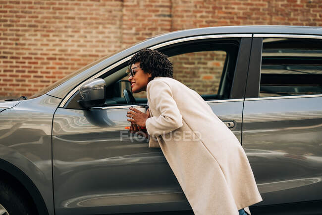 Vista lateral de la alegre hembra afroamericana sonriendo y mirando el espejo de un coche moderno de plata cerca del edificio de ladrillo - foto de stock