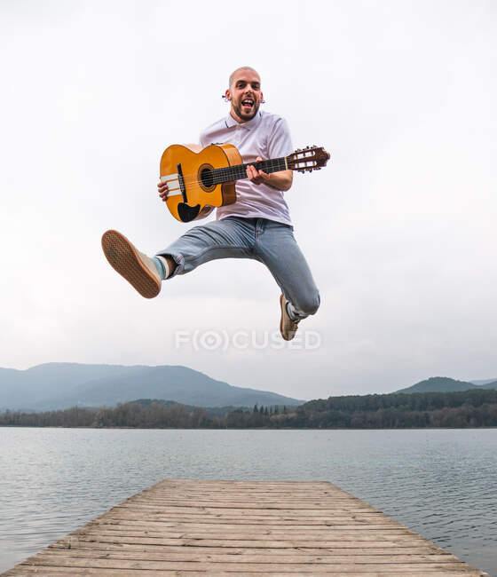 Full length of bearded guy in casual outfit jumping with guitar on wooden pier near lake with green mountains on background — Stock Photo
