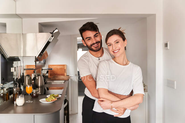 Side view of young romantic ethnic guy in white t shirt smiling and hugging happy girlfriend while looking at camera — Stock Photo