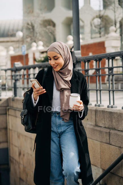 Délicieuse femme ethnique en hijab et vêtements à la mode marchant à l'étage avec boisson à emporter tout en surfant sur Internet sur smartphone et profiter week-end en ville — Photo de stock