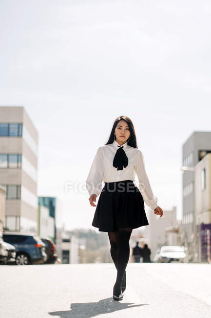 Низкий угол азиатской женщины в блузке и юбке, идущей по проезжей части к зданиям и смотрящей в камеру — стоковое фото