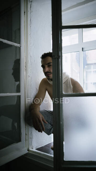 Hombre pensativo sentado de rodillas en el alféizar de la ventana en casa - foto de stock