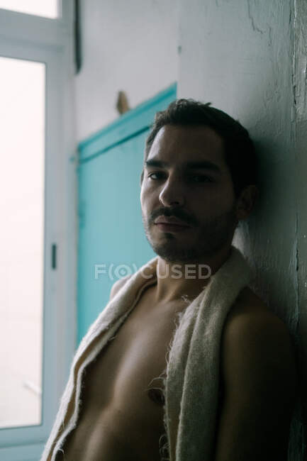 Спокойный мужчина без рубашки с бородой, опирающийся на потрепанную стену дома и смотрящий в камеру — стоковое фото