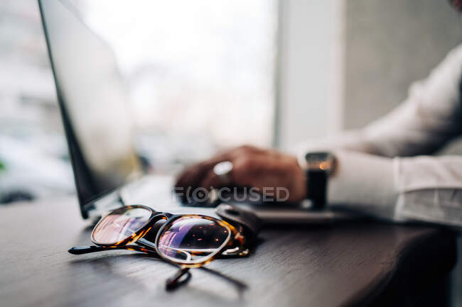 Crop anonymous male executive using portable computer at table with modern eyeglasses in cafe — Stock Photo