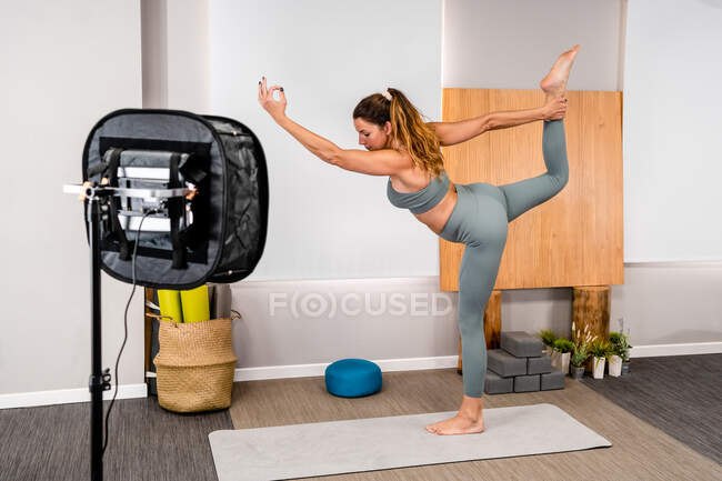 Side view of concentrated young female instructor in activewear standing in Lord of the Dance asana on mat while filming tutorial video during yoga practice — Stock Photo