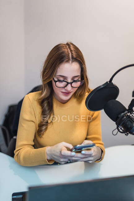 Весела жінка в окулярах текстові повідомлення на мобільний телефон, сидячи за столом з ноутбуком і мікрофоном під час роботи в студії мовлення — стокове фото