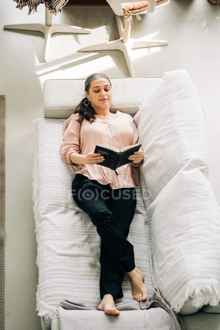 From above full body of barefoot female reading book while lying down on comfortable sofa in living room with green plant at home — Stock Photo